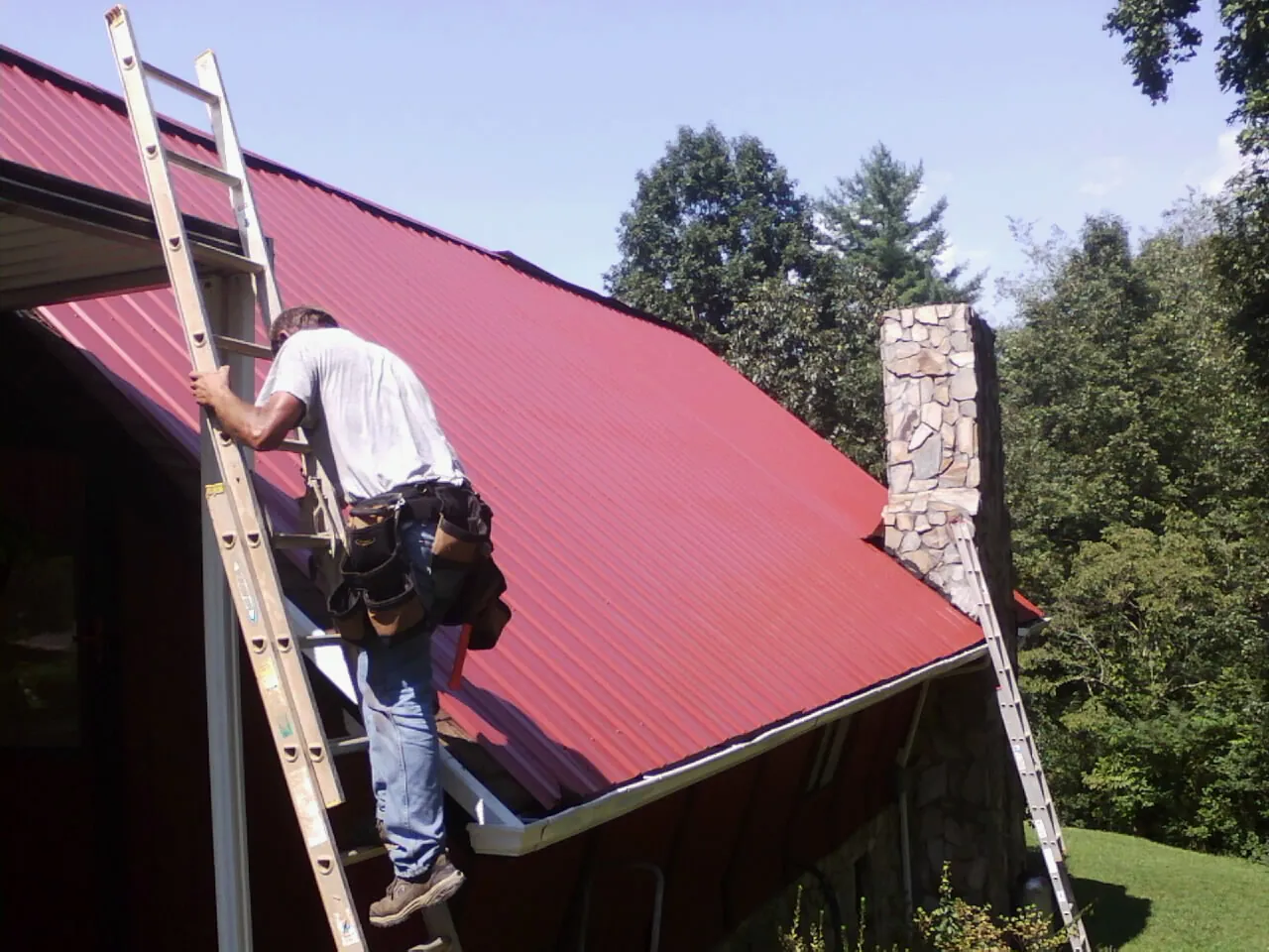 Roofer on a ladder beside a red metal roof and stone chimney — Soffit & Fascia Repair by Roofing Brothers in Riverdale